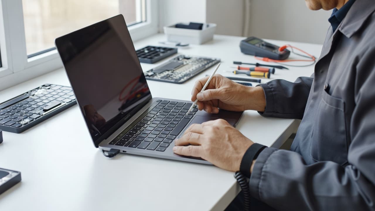 A technician wears an anti-static wrist strap while using precision tools to clean a MacBook keyboard. The bright workstation features a pristine desk surface illuminated by soft, natural window light.